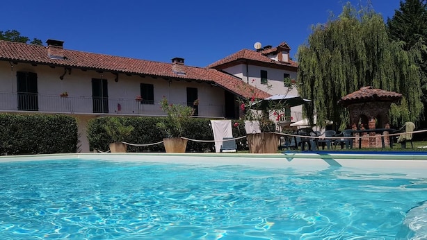 A clear swimming pool with a tiled deck area, surrounded by buildings with red-tiled roofs and white walls. - La Cascina del Castello (Asti)