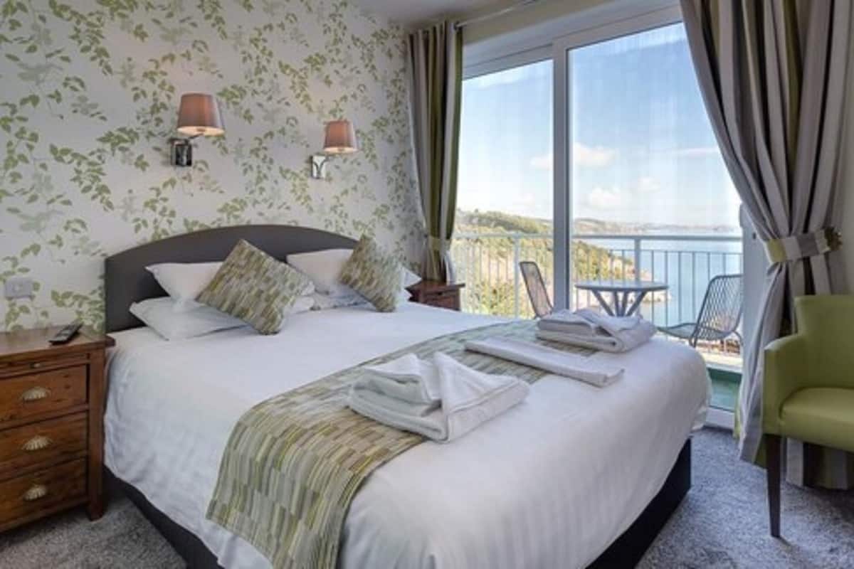 Quarto de hotel com uma cama grande, mesa de cabeceira, cadeira e varanda com vista.