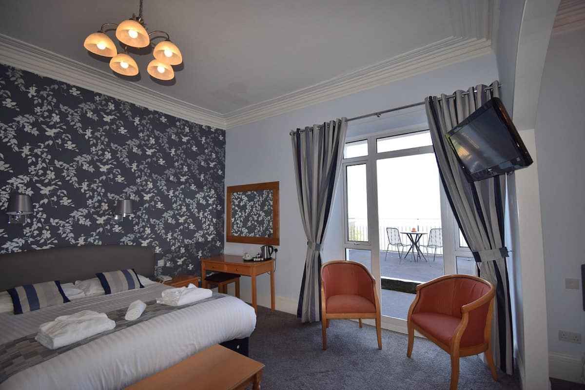 Quarto de hotel com cama, mesa de cabeceira, cadeira e televisão fixada na parede.