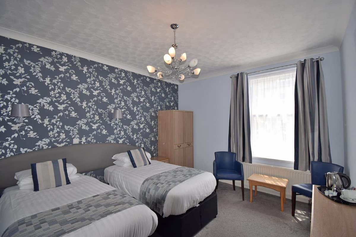 Quarto de hotel com duas camas, um candelabro, um guarda-roupa, uma secretária com uma cadeira e uma janela com cortinas.