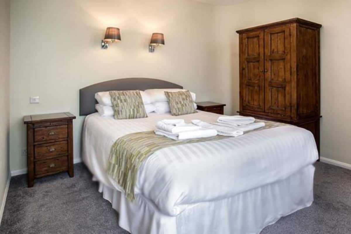 Uma cama bem arrumada com lençóis e almofadas brancos, um guarda-roupa de madeira e duas luminárias fixadas na parede.