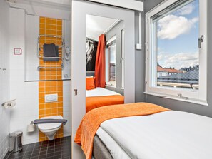 Habitación de hotel con baño que cuenta con azulejos naranjas, inodoro y zona de ducha. La habitación tiene dos camas con ropa de cama naranja y una ventana con vistas a la ciudad.