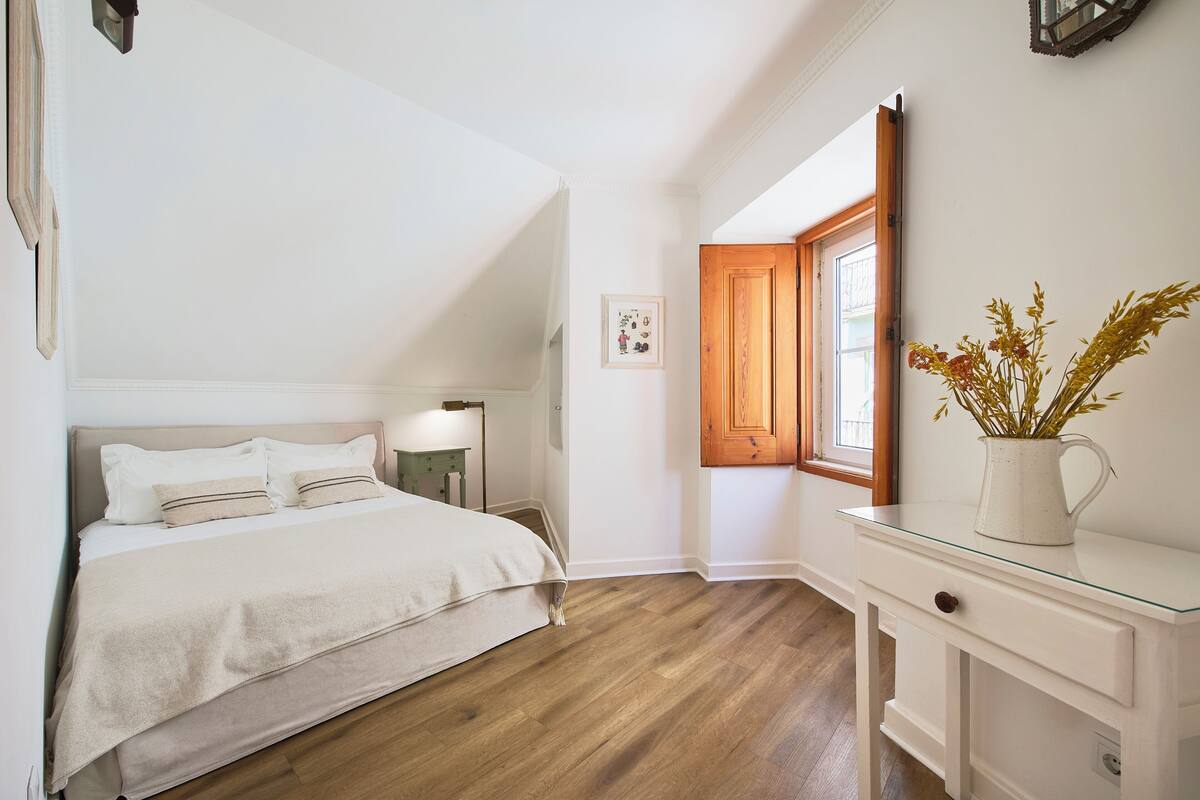 Une chambre à coucher comprenant un lit, une table de chevet, une commode, une fenêtre avec des volets en bois et un tableau accroché au mur.