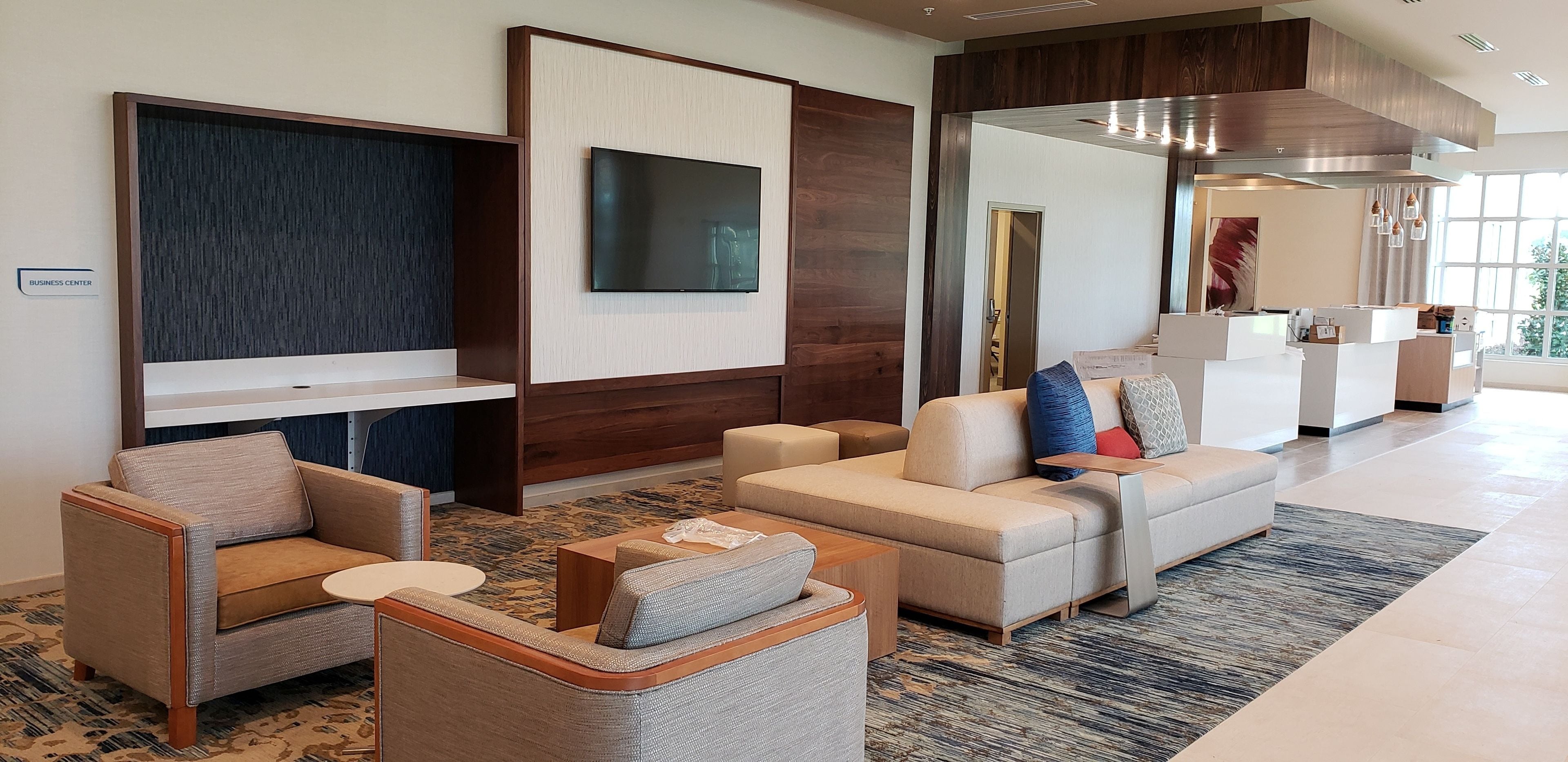 A modern lobby with a reception desk, seating area with sofas and armchairs, a large flat-screen TV, and a wooden accent wall.