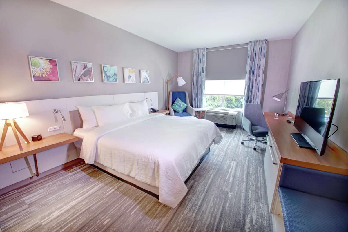 Habitación de hotel con una cama grande, un escritorio con computadora, un televisor y una ventana con cortinas.