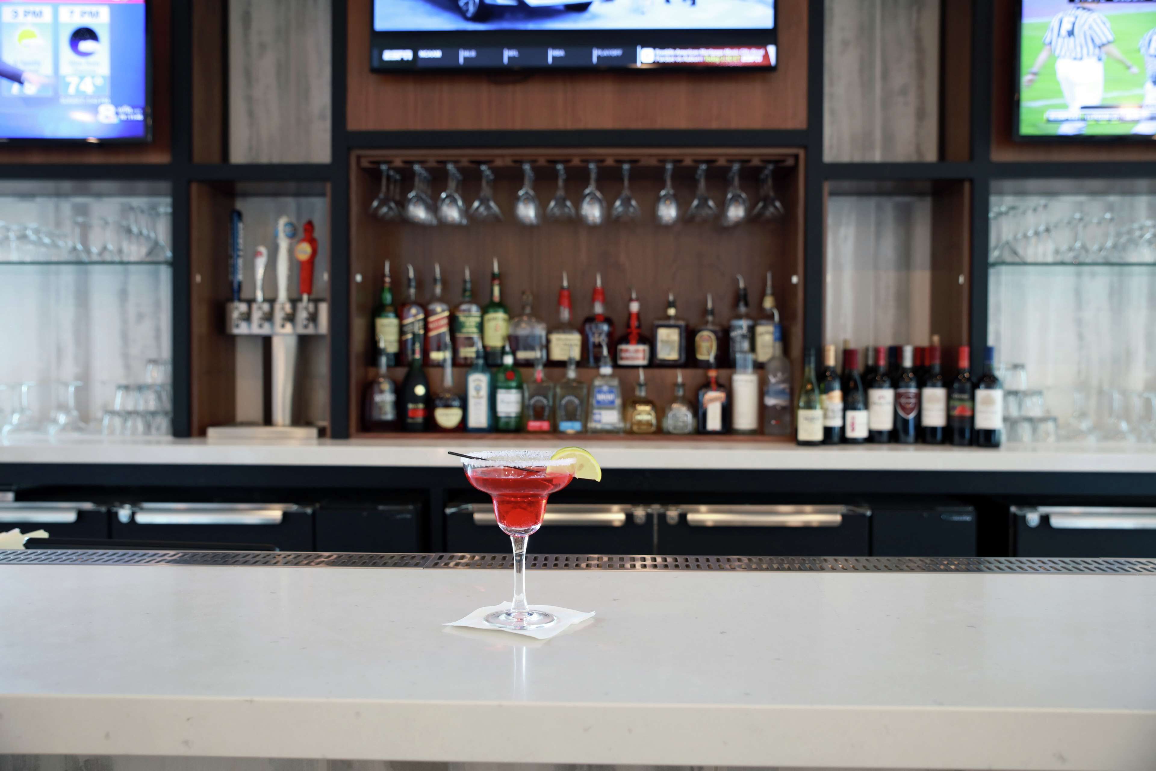A bar area with a martini glass on a bar counter, a television mounted above the bar, and shelves with various bottles and glasses in the background.