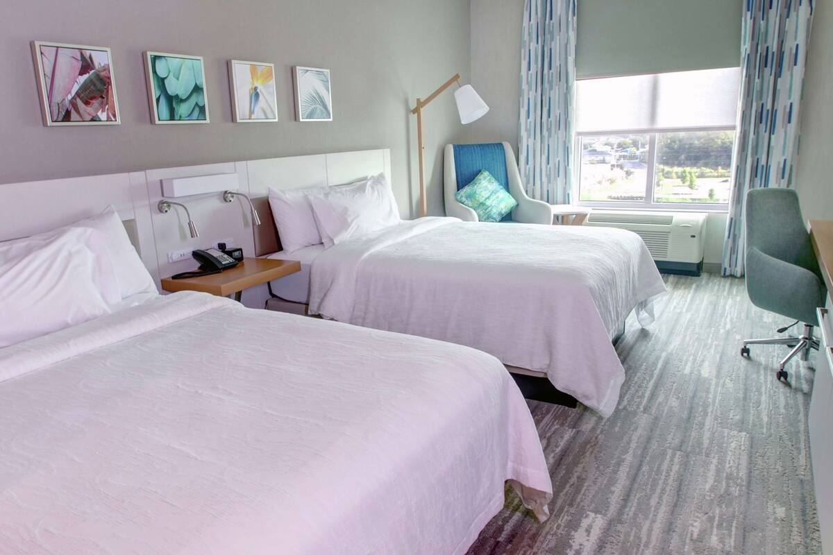 Habitación de hotel con dos camas, un escritorio con lámpara, una silla y una ventana con cortinas.