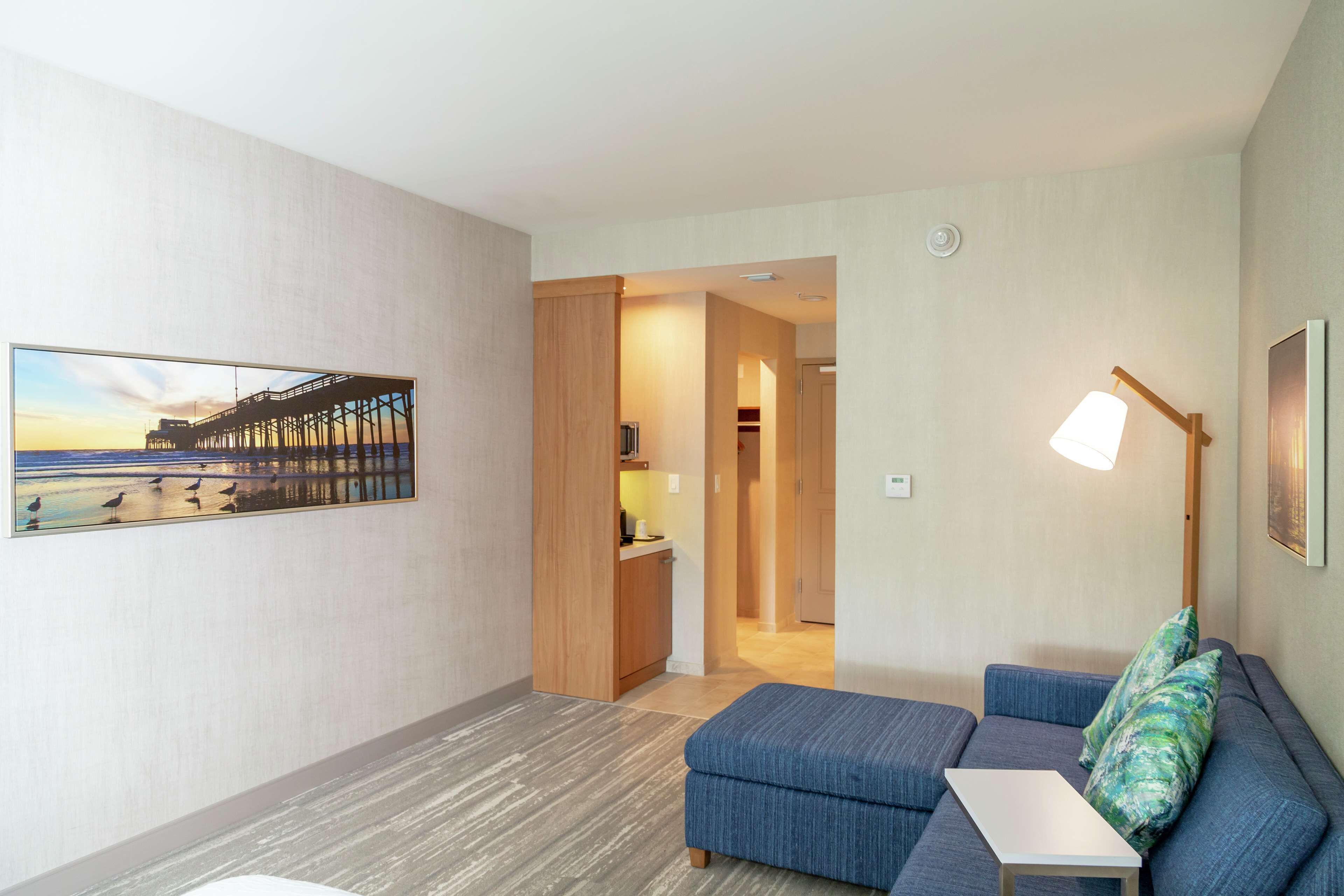 Ein modernes Hotelzimmer mit Holzboden, einem blauen Sofa, einem kleinen Tisch, einer Lampe und einem Gemälde eines Piers.