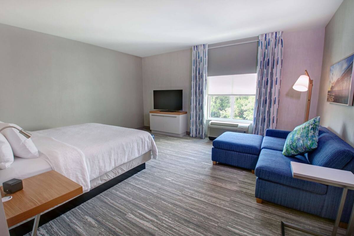 Una habitación de hotel con cama, sofá, televisión y ventana con cortinas.