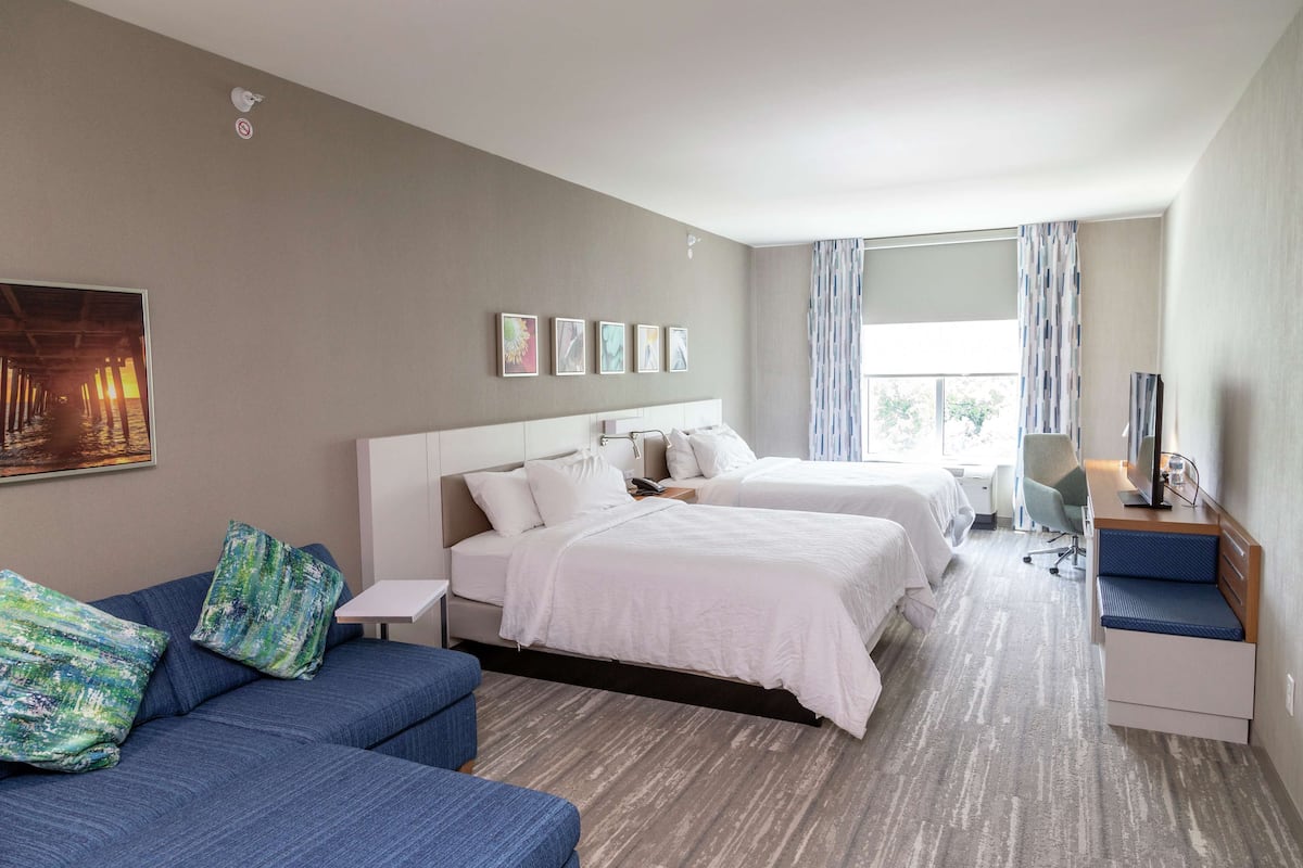 Habitación de hotel con dos camas, un sofá, un escritorio con silla y un amplio ventanal con cortinas.