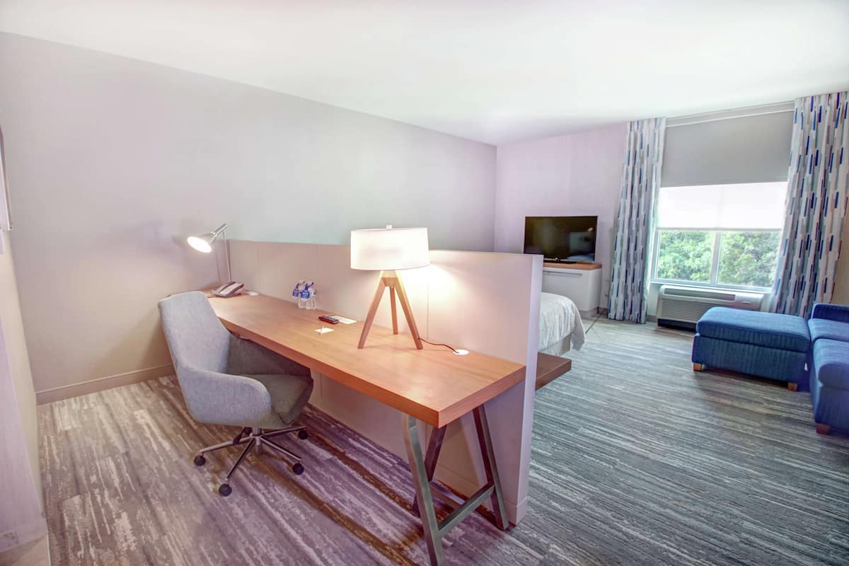 Una habitación de hotel con un escritorio de madera, una silla, una lámpara, un televisor, una cama y una ventana con cortinas.