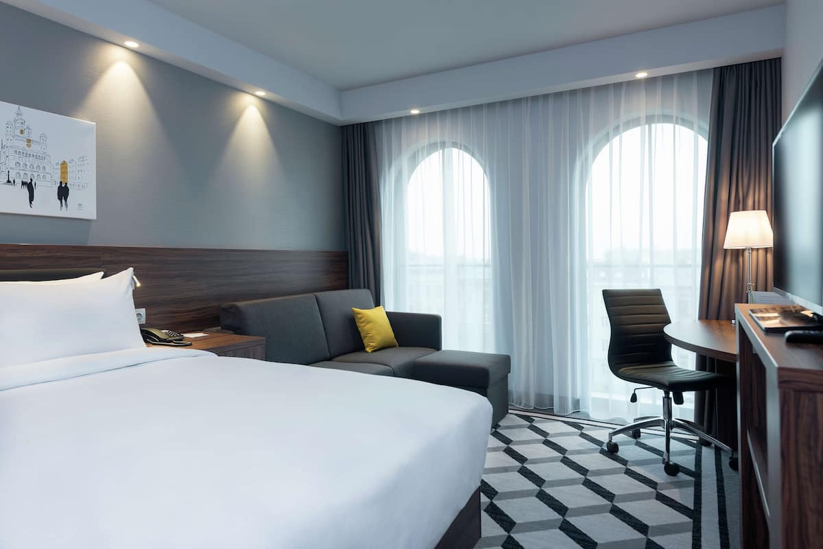 Une chambre d’hôtel avec un grand lit, un canapé, un bureau et une chaise. Il y a deux fenêtres avec des rideaux, un tableau au mur et une lampe sur le bureau.