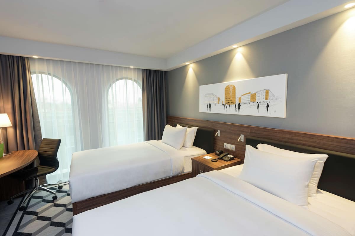 Une chambre d’hôtel avec deux lits, un bureau et une chaise. Il y a un tableau au mur et une fenêtre avec des rideaux.