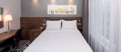 A hotel room with a large bed, a desk with a chair, a lamp, a phone, and a painting on the wall.