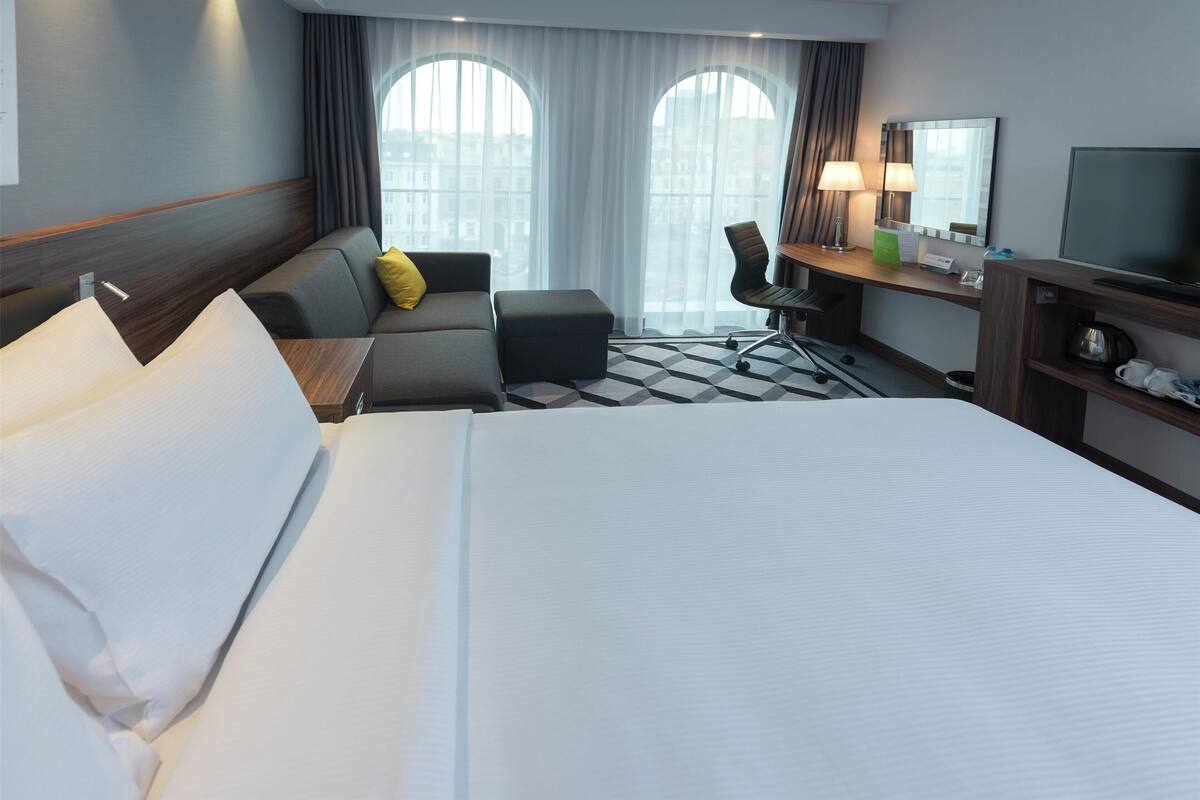 Une chambre d’hôtel avec un grand lit, un canapé avec un coussin jaune, un bureau avec un ordinateur et des lampes, et une télévision.