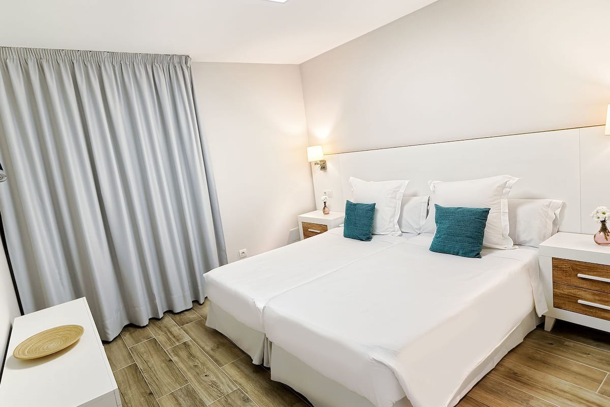 A hotel room with a large bed, two bedside tables, a nightstand, a wall-mounted lamp, and a window with curtains.