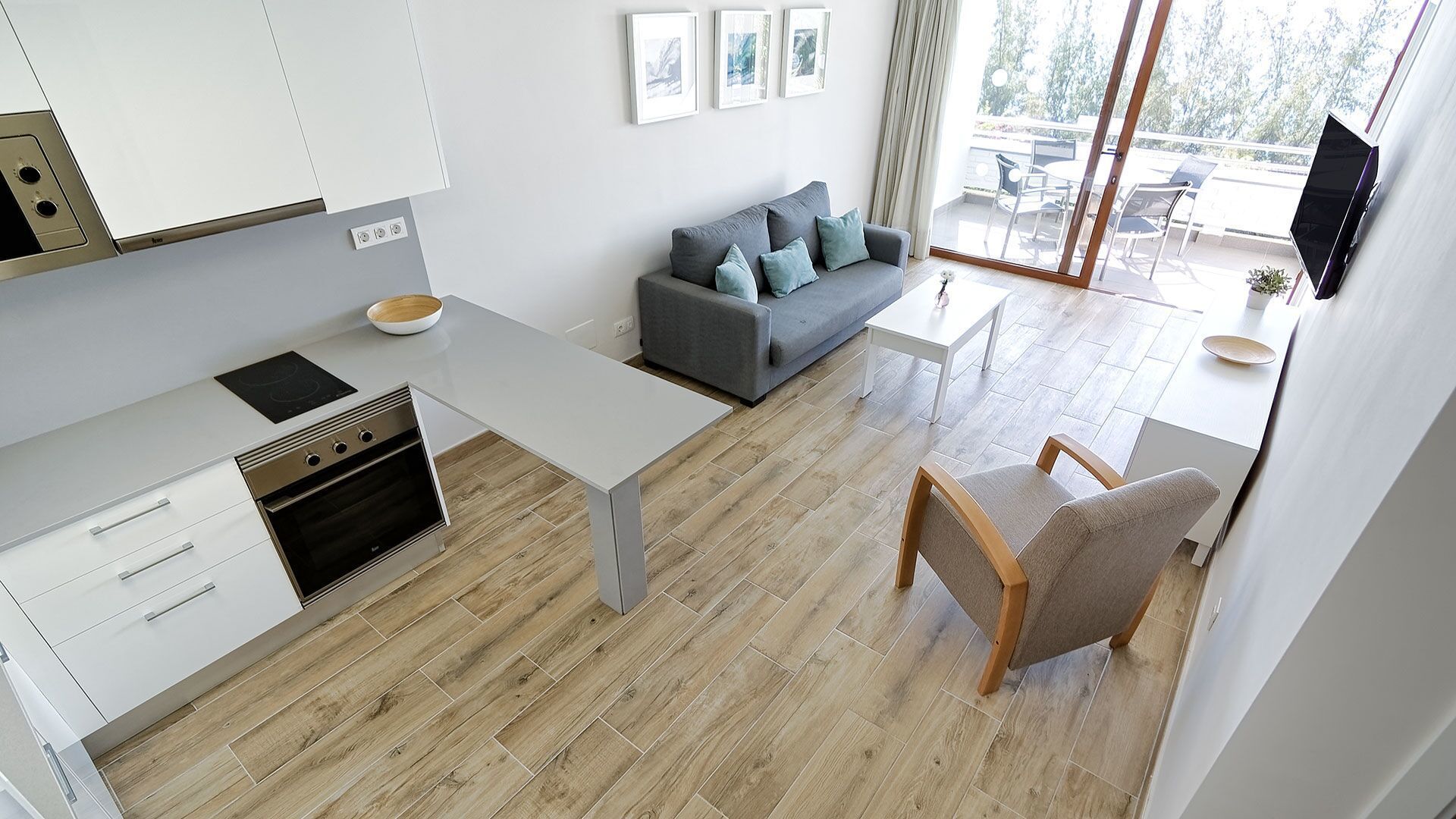 Eine moderne Küche mit Einbauofen, ein Essbereich mit Tisch und Stühlen, ein graues Sofa und ein an der Wand befestigter Fernseher.