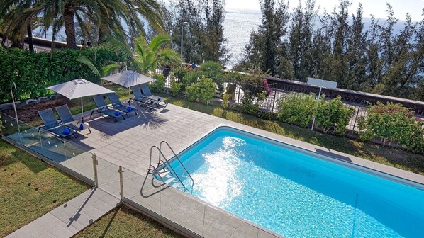 A rectangular swimming pool with a clear blue water surface, surrounded by a tiled deck area with lounge chairs and umbrellas.