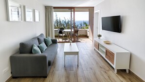 Een moderne woonkamer met een grijze bank, een witte salontafel en een flatscreen-televisie aan de muur. Er is een schuifdeur die leidt naar een terras buiten, met een tafel en stoelen.