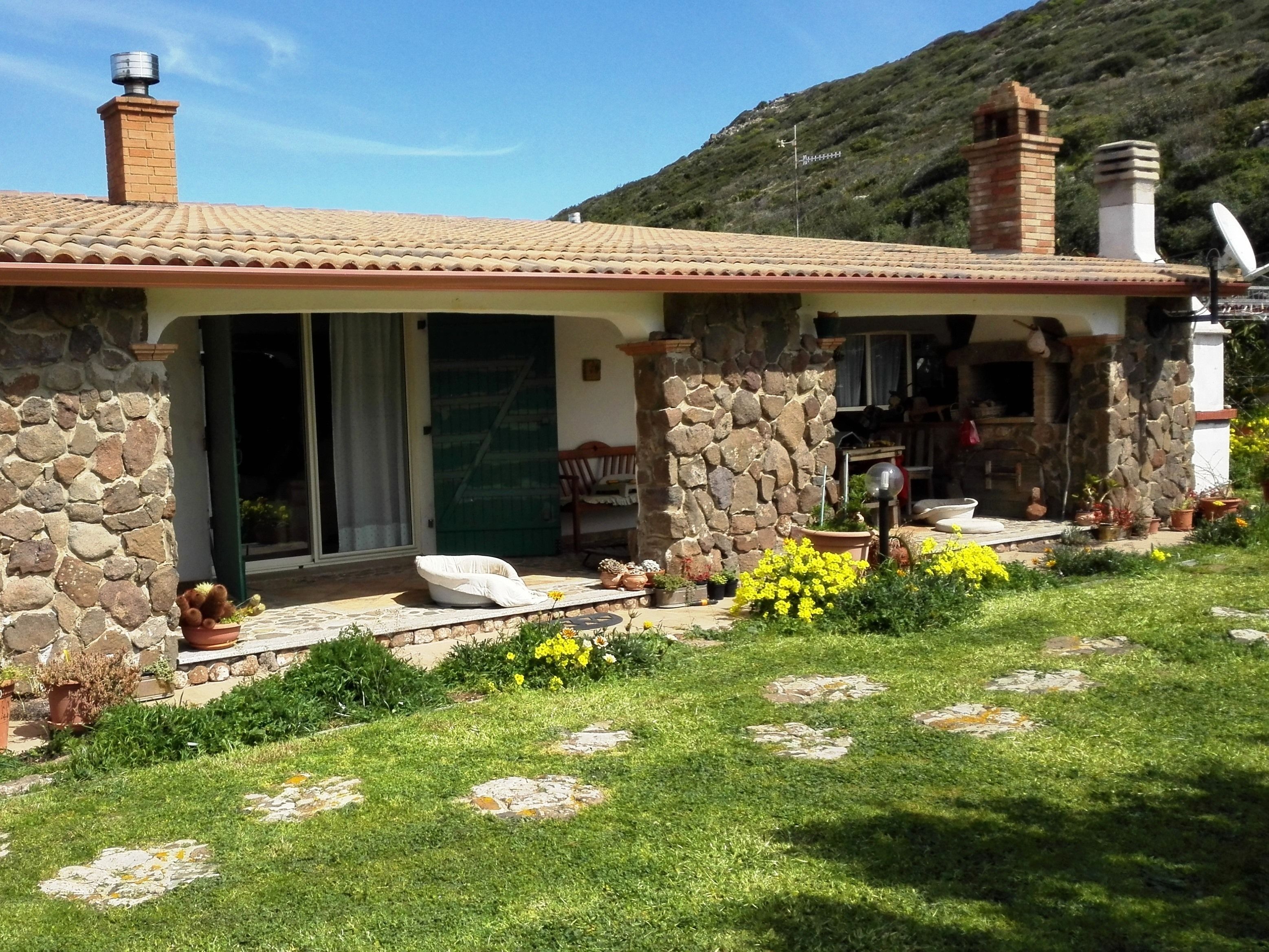A stone house with a tiled roof, a stone chimney, and a garden with yellow flowers.