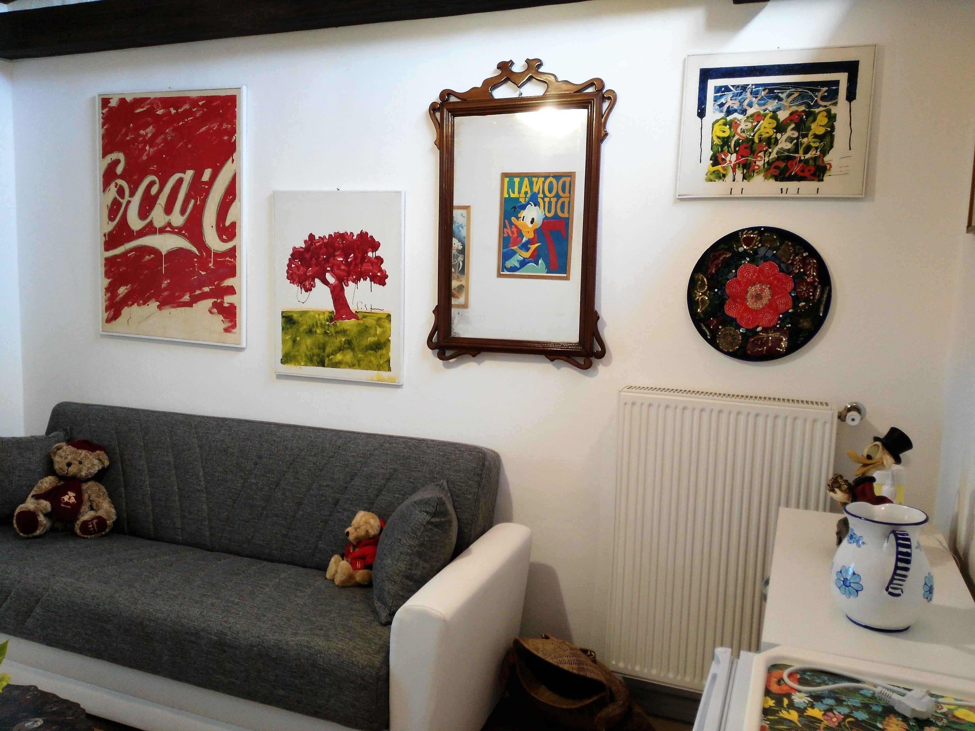 A living room with a grey sofa, two teddy bears, a framed picture of a tree, a framed picture of Donald Duck, a framed picture of a colorful abstract, and a decorative plate on the wall.