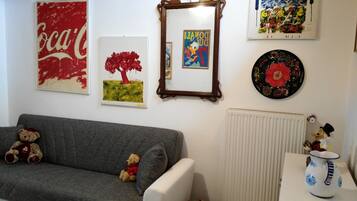 A living room with a grey sofa, two teddy bears, a framed picture of a tree, a framed picture of Donald Duck, a framed picture of a colorful abstract, and a decorative plate on the wall.