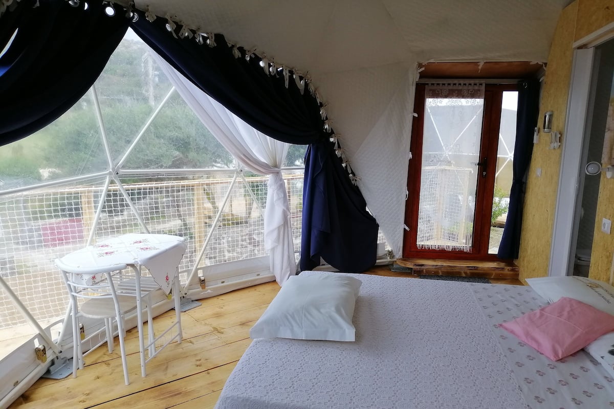 A room with a bed, a table, and a chair. There is a window with curtains, and a door leading outside.