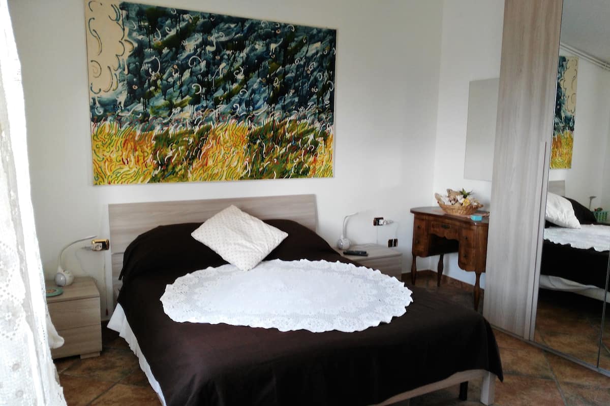 A bedroom with a bed, a nightstand, a bedside table, a mirror, and a painting on the wall.