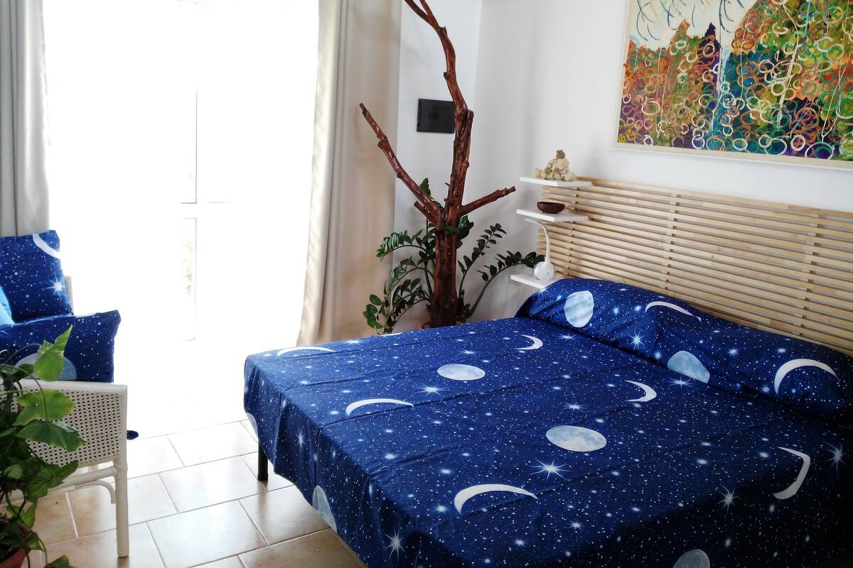 A bedroom with a bed covered in a starry night theme, a potted plant, a white chair, and a painting on the wall.
