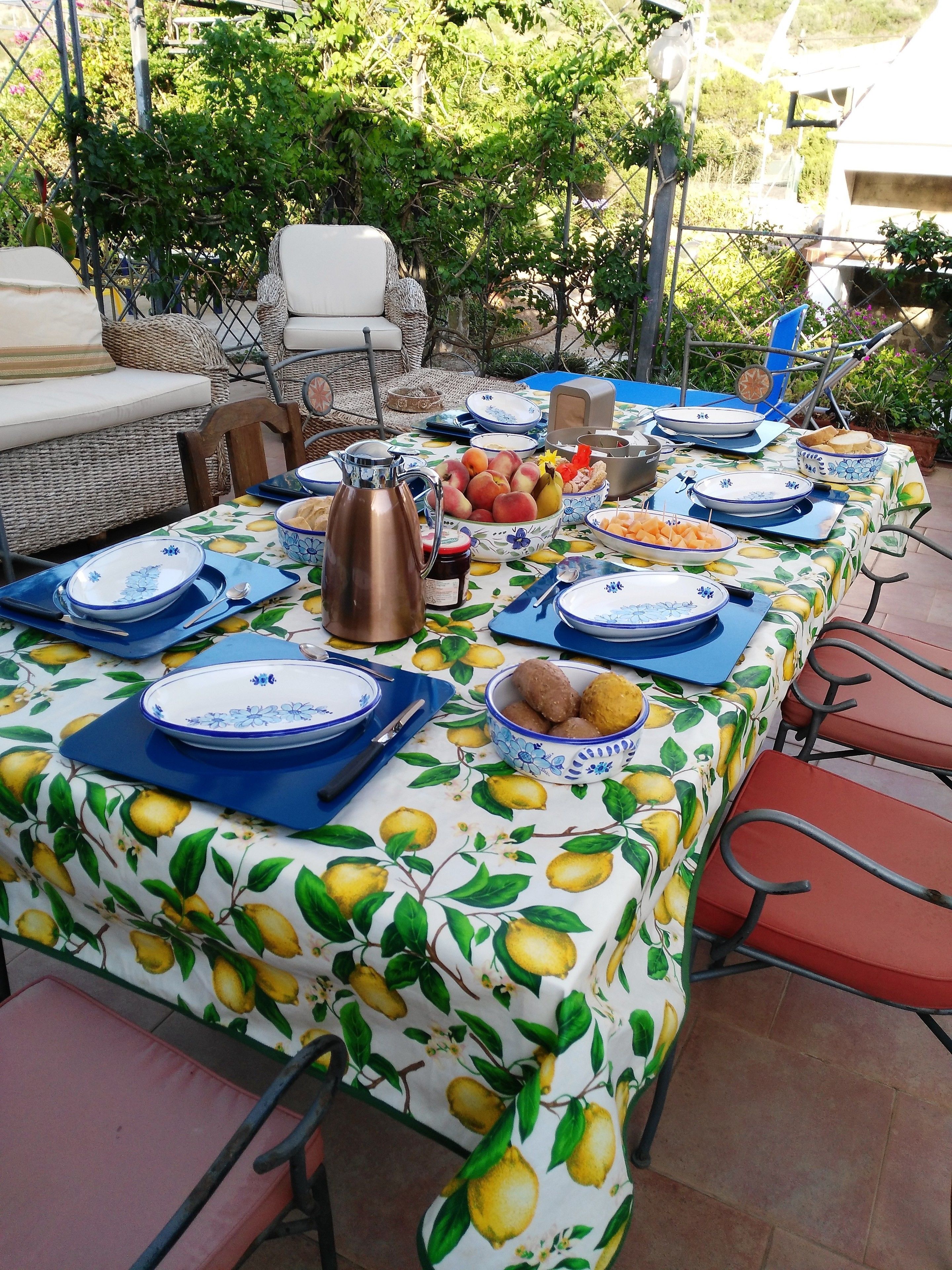 A table set outdoors with a tablecloth featuring a lemon pattern, surrounded by chairs.