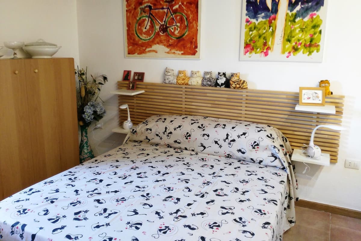 A bedroom with a bed, a wooden headboard, a bedside table with a lamp, and a wall with paintings.