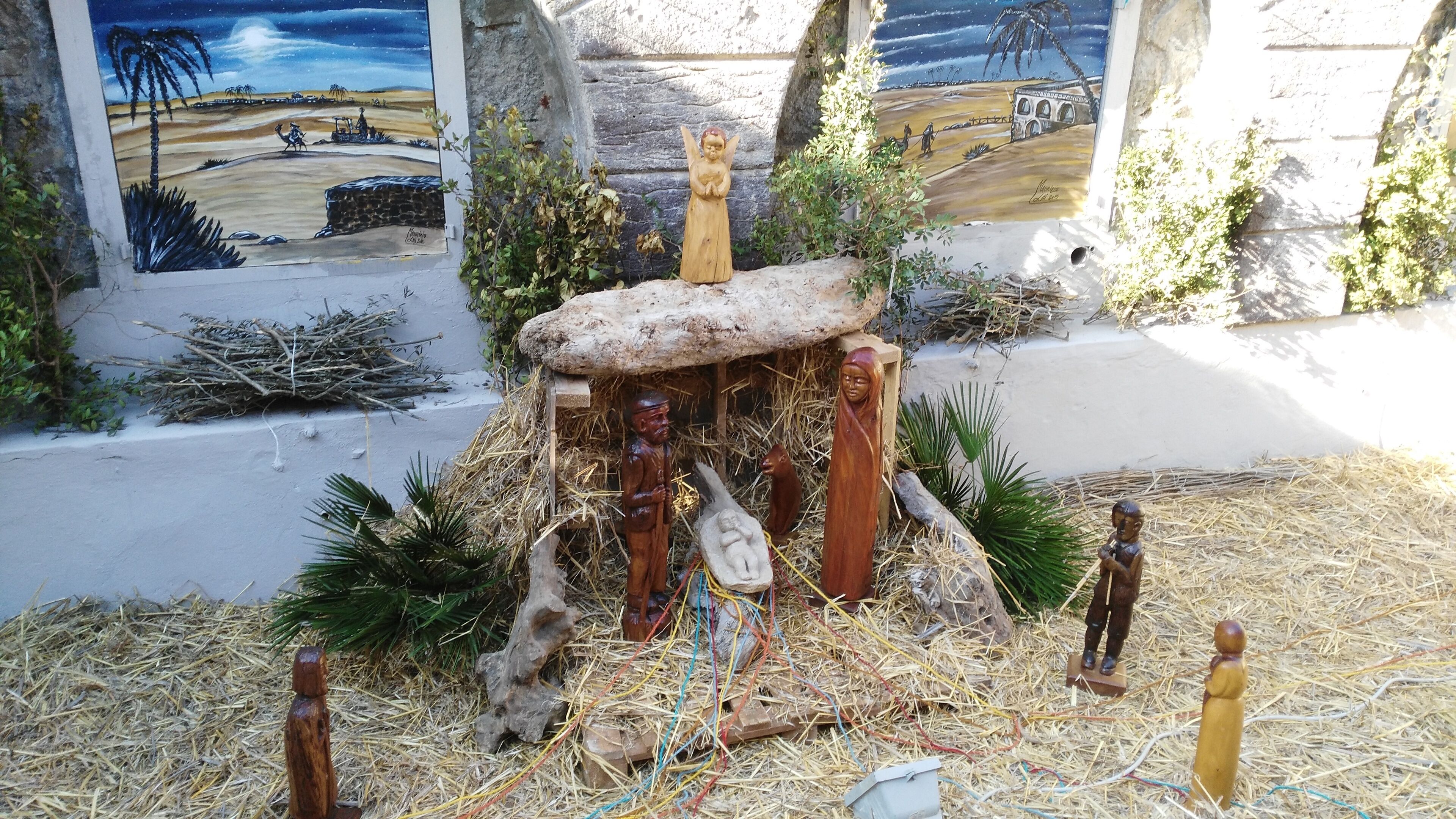 A nativity scene with a stable, figures of Mary, Joseph, baby Jesus, and animals, set against a backdrop of a mural depicting a desert landscape.