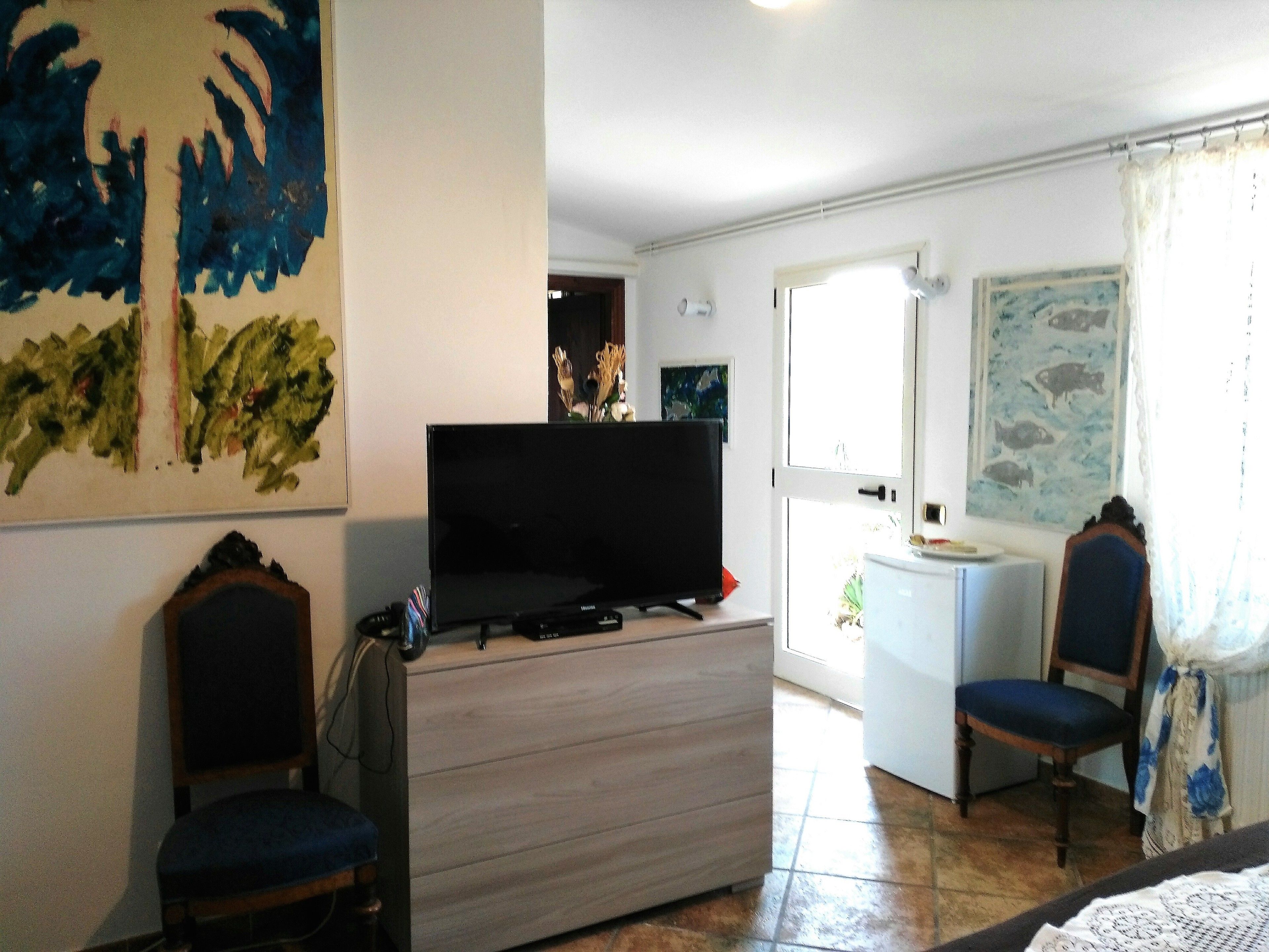A living room with a flat-screen TV on a wooden cabinet, two chairs, a small table, and artwork on the walls.
