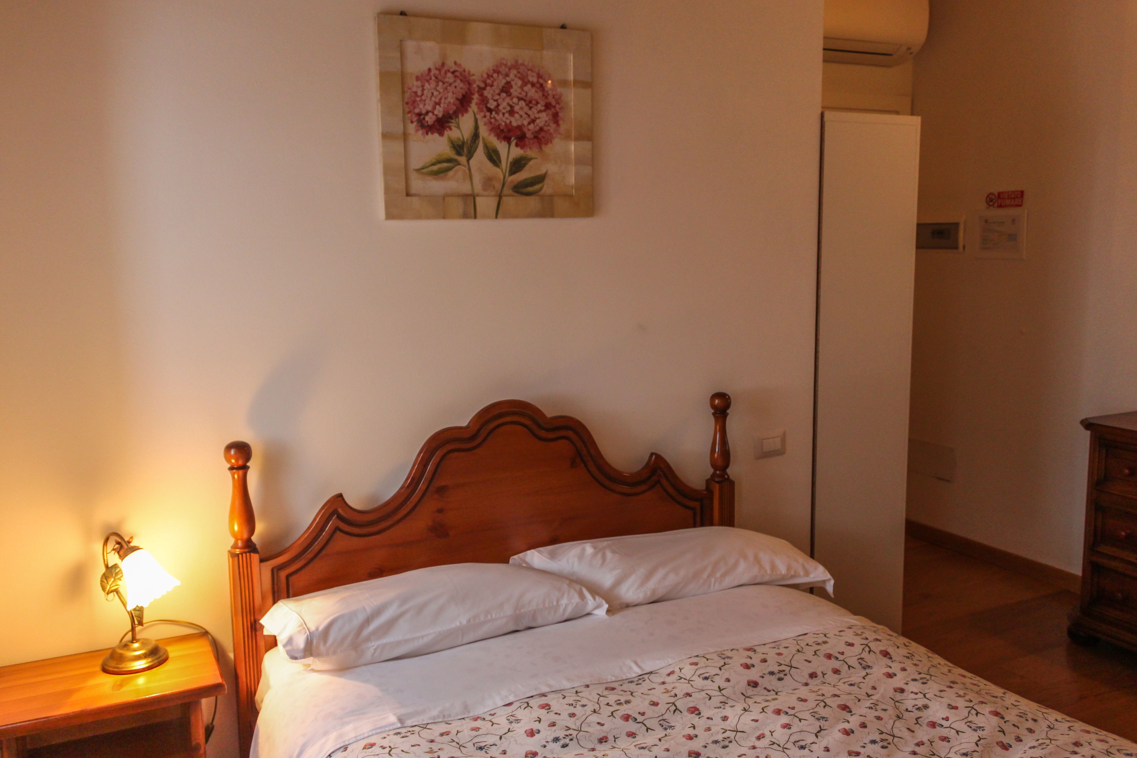 Un dormitorio con una cama de madera, ropa de cama blanca, una lámpara de noche y una obra de arte floral en la pared.