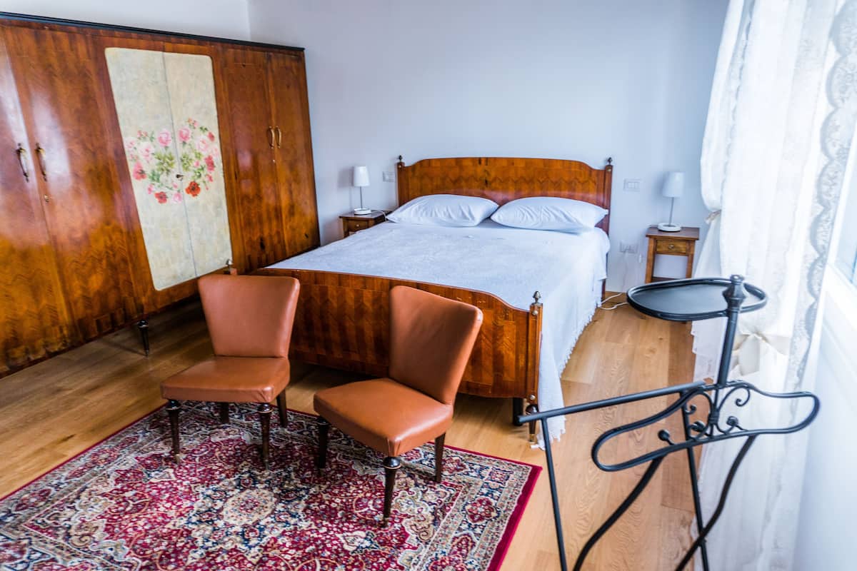 Een slaapkamer met een houten kledingkast, een bed met witte lakens, twee bruine stoelen, een getextureerd vloerkleed en een klein tafeltje.