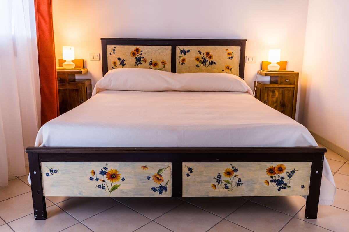 Een bed met een bloemrijk hoofdeinde en voeteinde, twee nachtkastjes met lampen, en een gordijn.