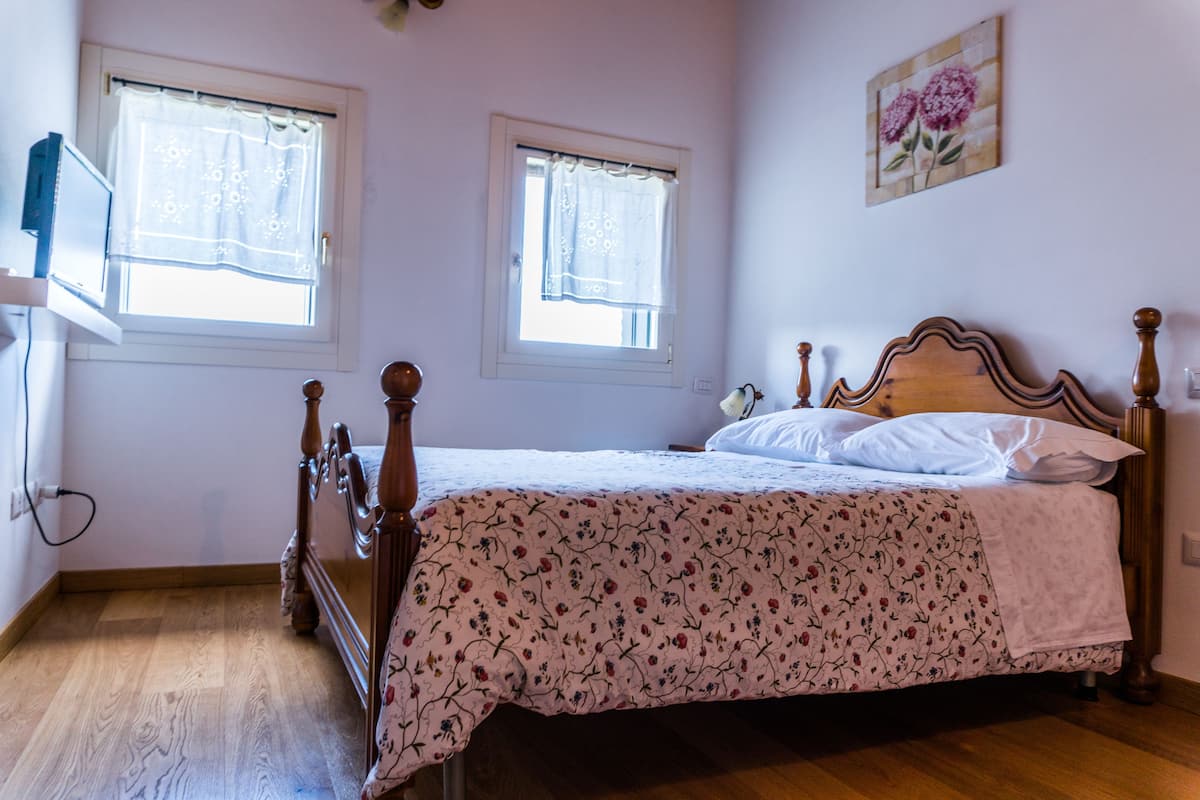 Een slaapkamer met een houten bed, twee ramen met dunne gordijnen, een schilderij aan de muur en een houten vloer.