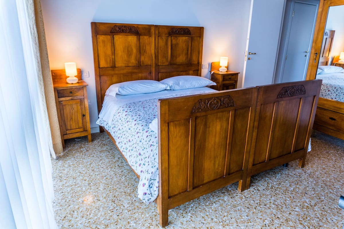 Een houten bed met een bloemenbedekking, twee nachtkastjes met lampen en een houten garderobe.