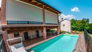 A swimming pool with a wooden deck, a covered seating area, and a view of a white building with a brick facade.