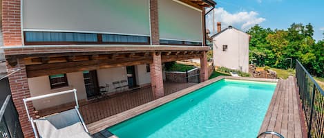 A swimming pool with a wooden deck, a covered seating area, and a view of a white building with a brick facade.