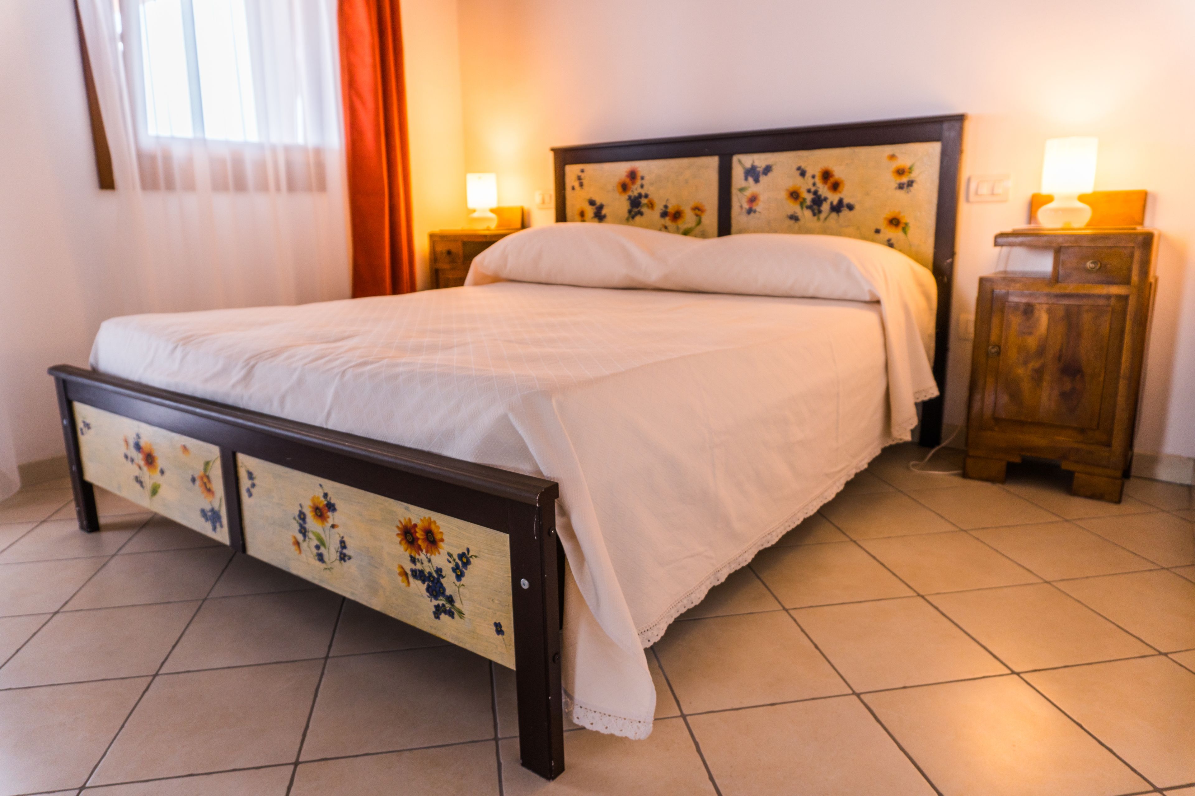 A neatly made bed with a floral headboard, a bedside table with a lamp, and a wooden nightstand with a lamp.