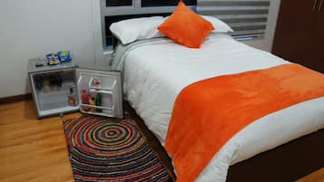 Uma cama bem arrumada com um cobertor e travesseiro laranja, uma pequena geladeira com lanches, e um tapete colorido sobre piso de madeira.
