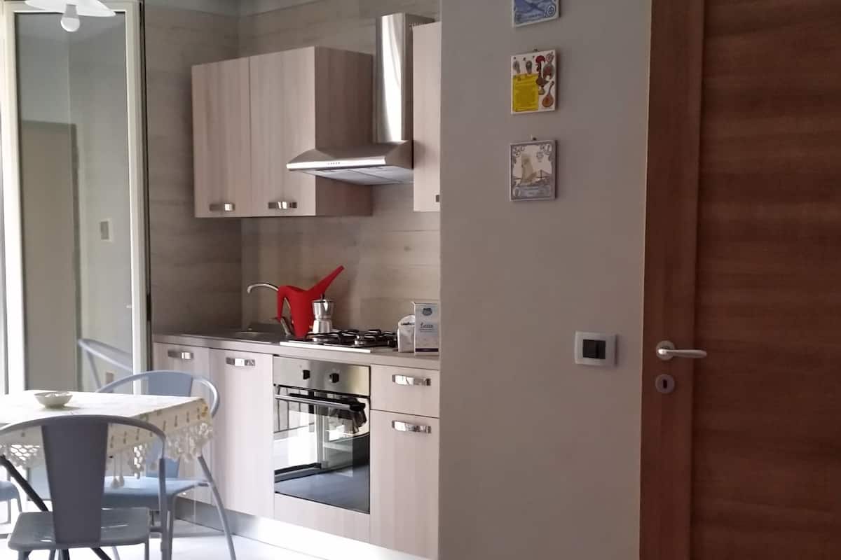 A room with two beds, a kitchen area with cabinets and a stove, and a dining table with chairs.