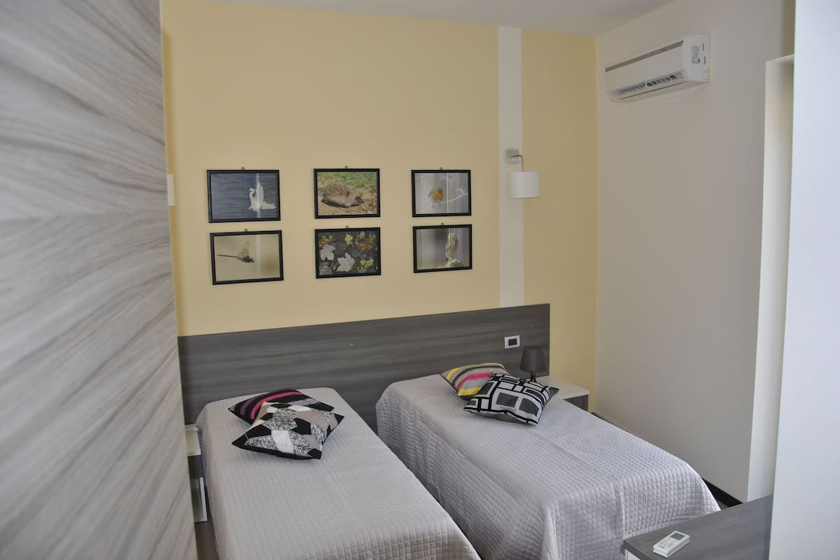 A hotel room with two single beds, a wall-mounted air conditioner, and four framed pictures.