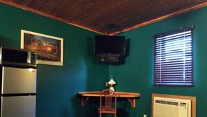 A room with teal walls, a wooden desk, a chair, a television mounted on the wall, a window with blinds, a microwave, and a radiator. - Comfort Zone Inn (Rothsay)