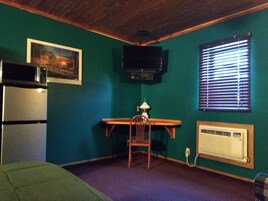 A room with teal walls, a wooden desk, a chair, a television mounted on the wall, a window with blinds, a microwave, and a radiator.