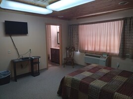A bedroom with a bed, a desk with a computer, a TV mounted on the wall, and a window with curtains.