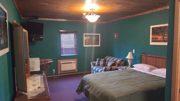 A bedroom with a bed, a television, a window with blinds, a heater, and a ceiling fan. - Comfort Zone Inn (Rothsay)