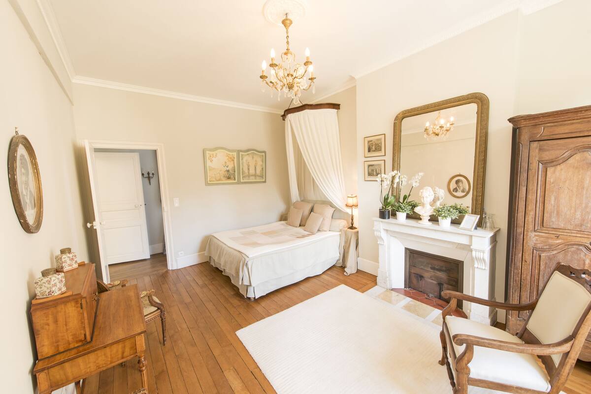 A bedroom with a bed, a chair, a fireplace, and a mirror.