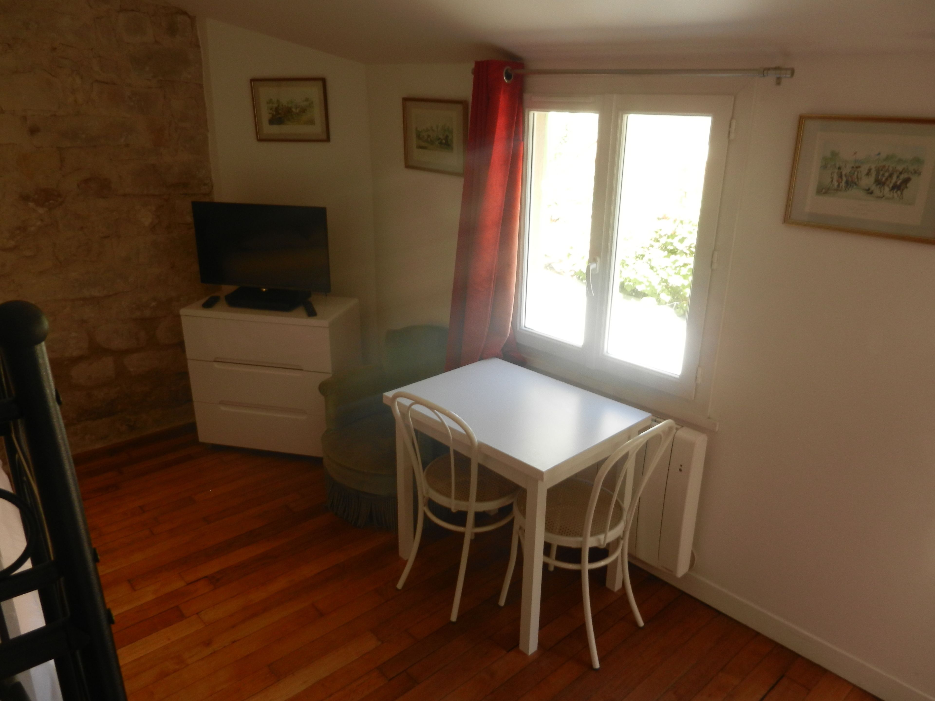 Una habitación pequeña con una mesa y sillas blancas, un televisor sobre un aparador y una ventana con cortina roja.